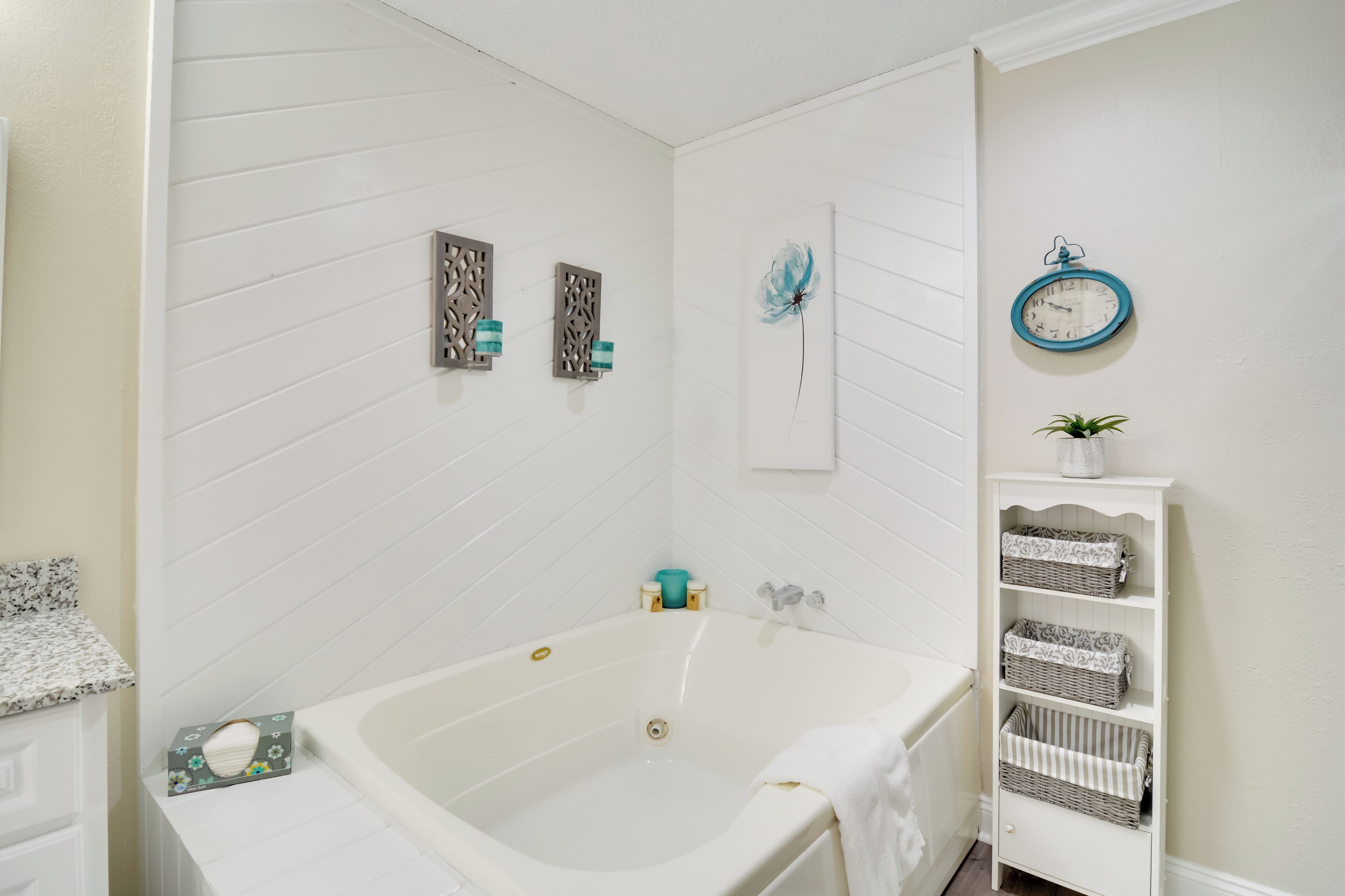 A bathroom with a white bathtub, a white shelf with baskets, a clock, and decorative wall panels.