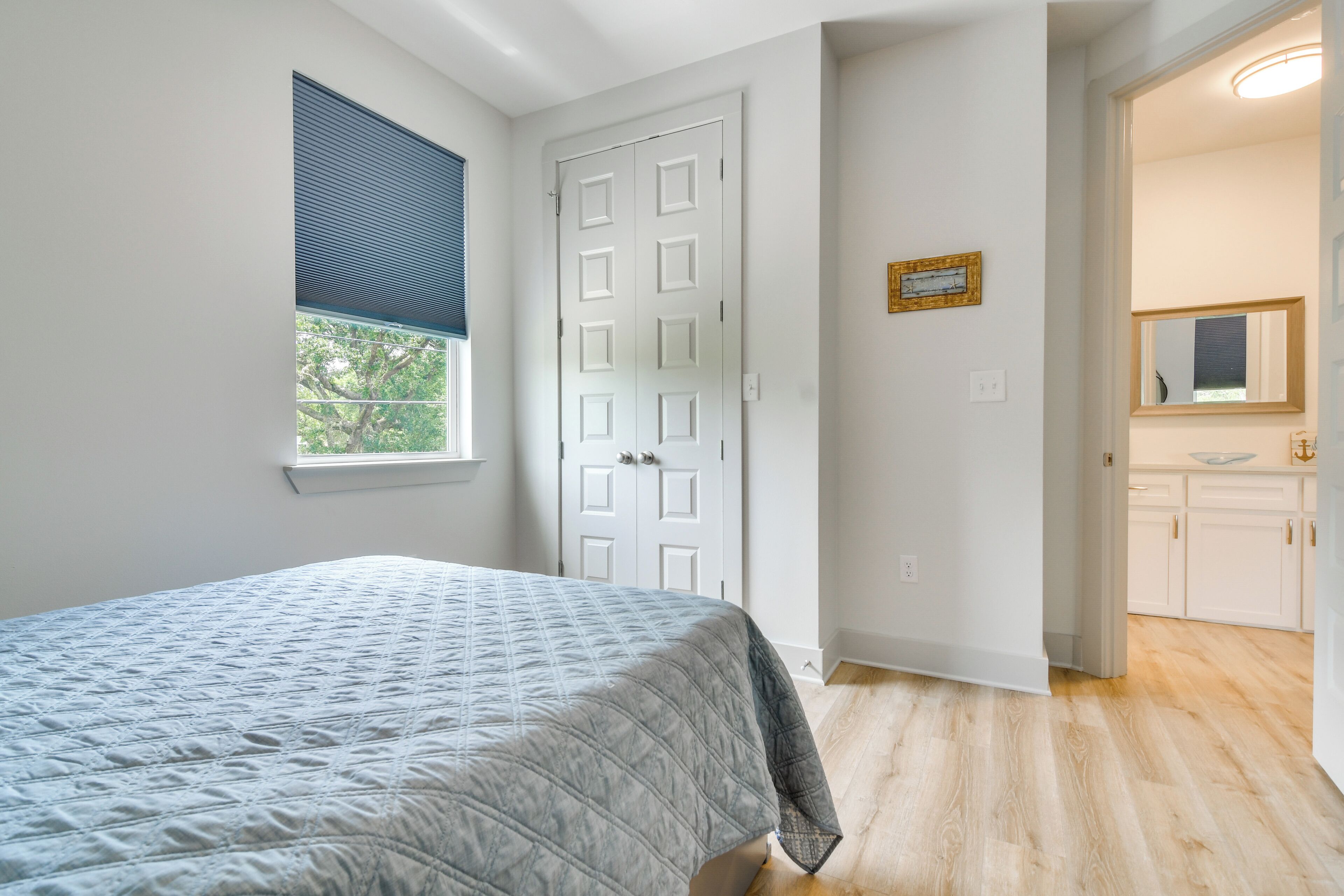A bedroom with a bed, a window with blinds, a white door, and a bathroom visible through an open door.