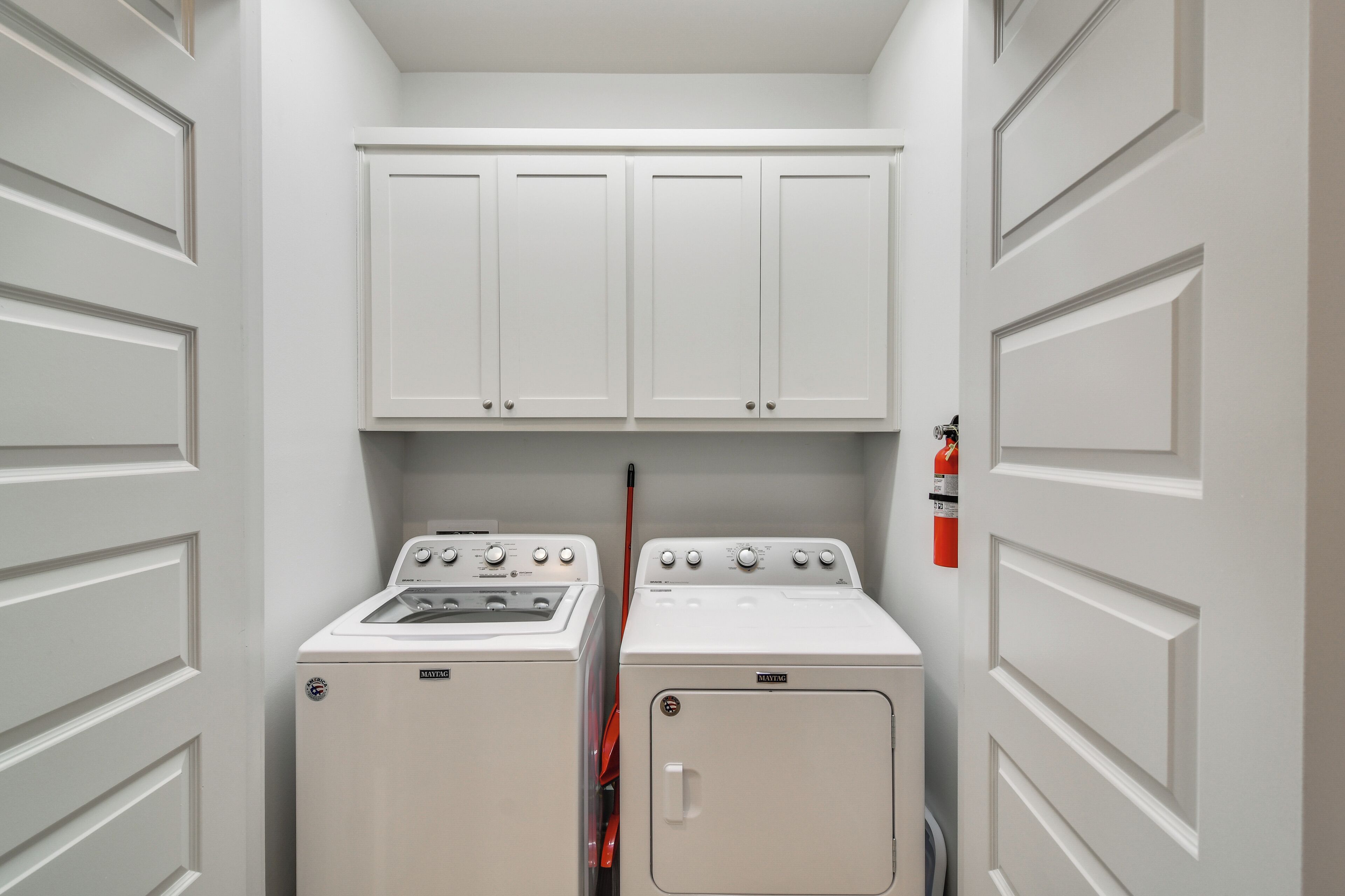 A laundry room with two washing machines and a dryer, white cabinets above, and a fire extinguisher on the wall.