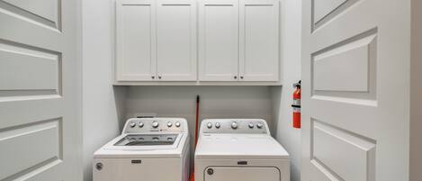 A laundry room with two washing machines and a dryer, white cabinets above, and a fire extinguisher on the wall.