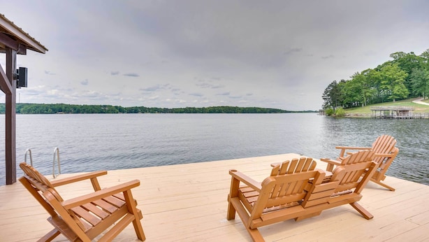Um deque de madeira com cadeiras Adirondack com vista para um lago, um píer e uma colina arborizada.