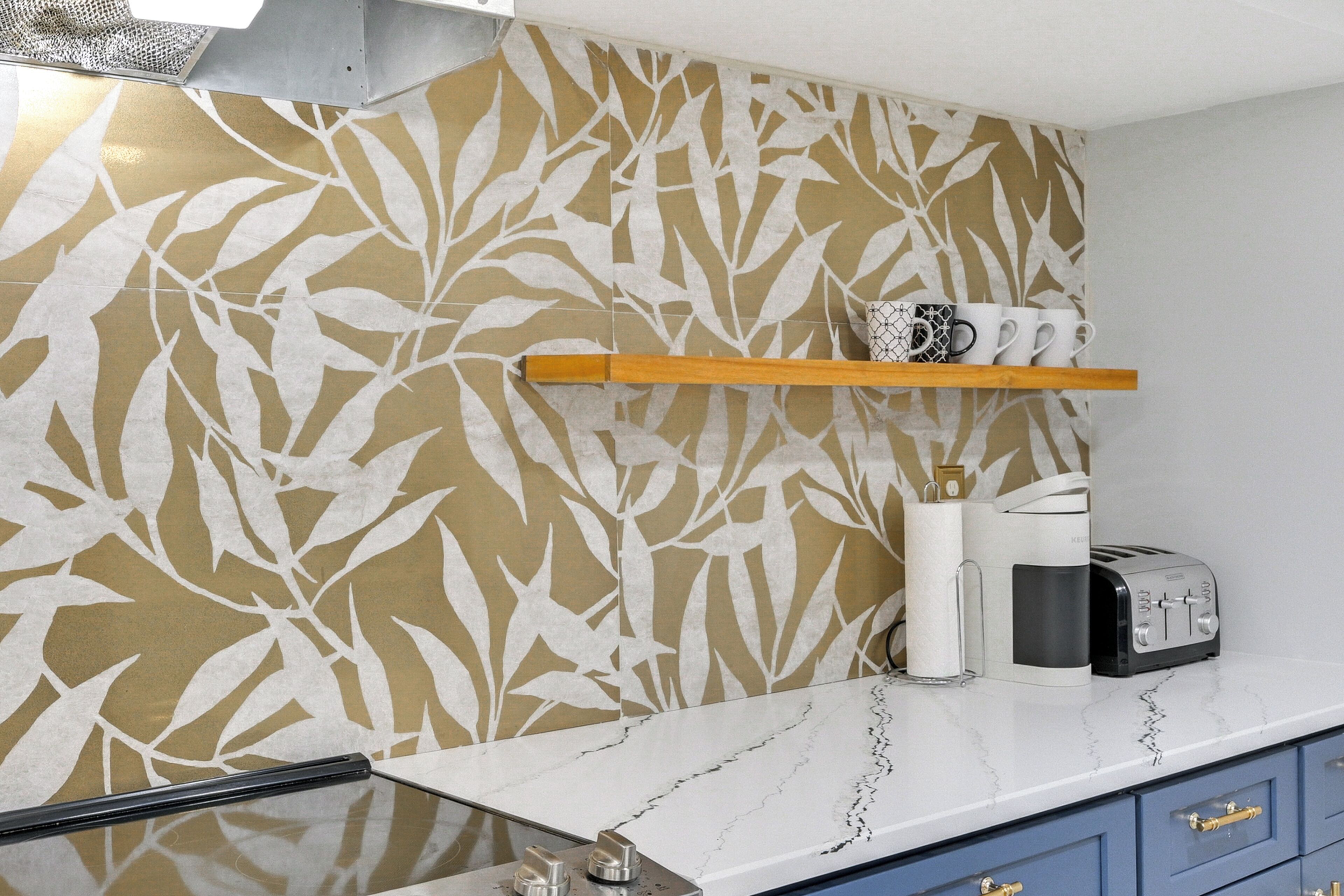 A kitchen with a leaf-patterned backsplash, a wooden shelf with mugs, a coffee maker, and a toaster.