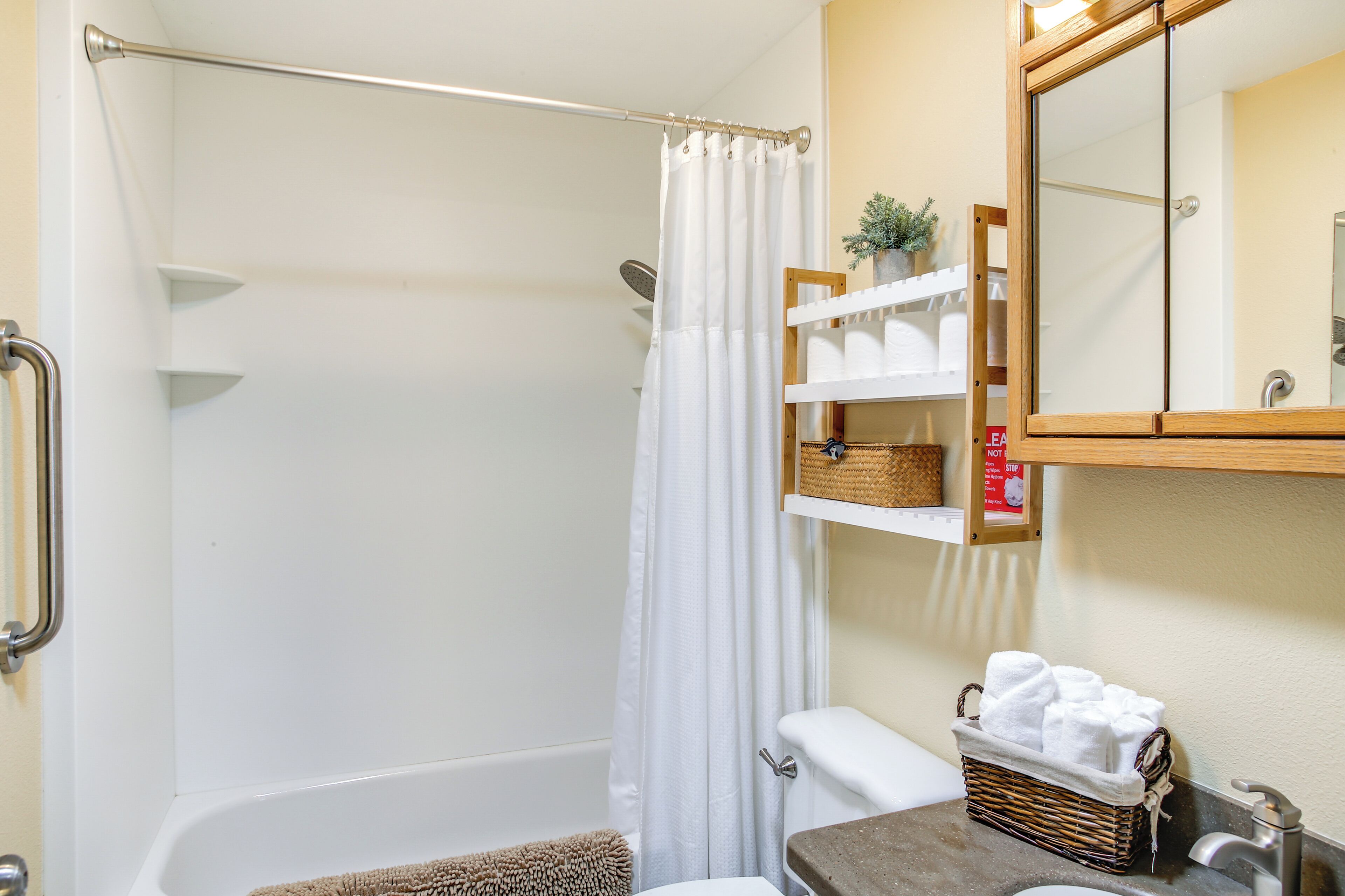 A bathroom with a bathtub, a shower curtain, a toilet, a sink with a mirror, and a shelf with towels and a small plant.