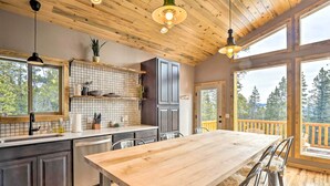 A modern kitchen with a wooden table and bar stools, large windows, and a view of trees. - Mountainside Fairplay Cabin w/ Game Room & Sauna! (Fairplay)
