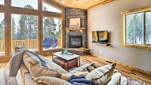 A cozy living room with a stone fireplace, large windows, and a TV mounted on the wall. - Mountainside Fairplay Cabin w/ Game Room & Sauna! (Fairplay)