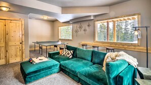 A cozy room with a green sectional sofa, a ping pong table, and a large window with a view of trees. - Mountainside Fairplay Cabin w/ Game Room & Sauna! (Fairplay)