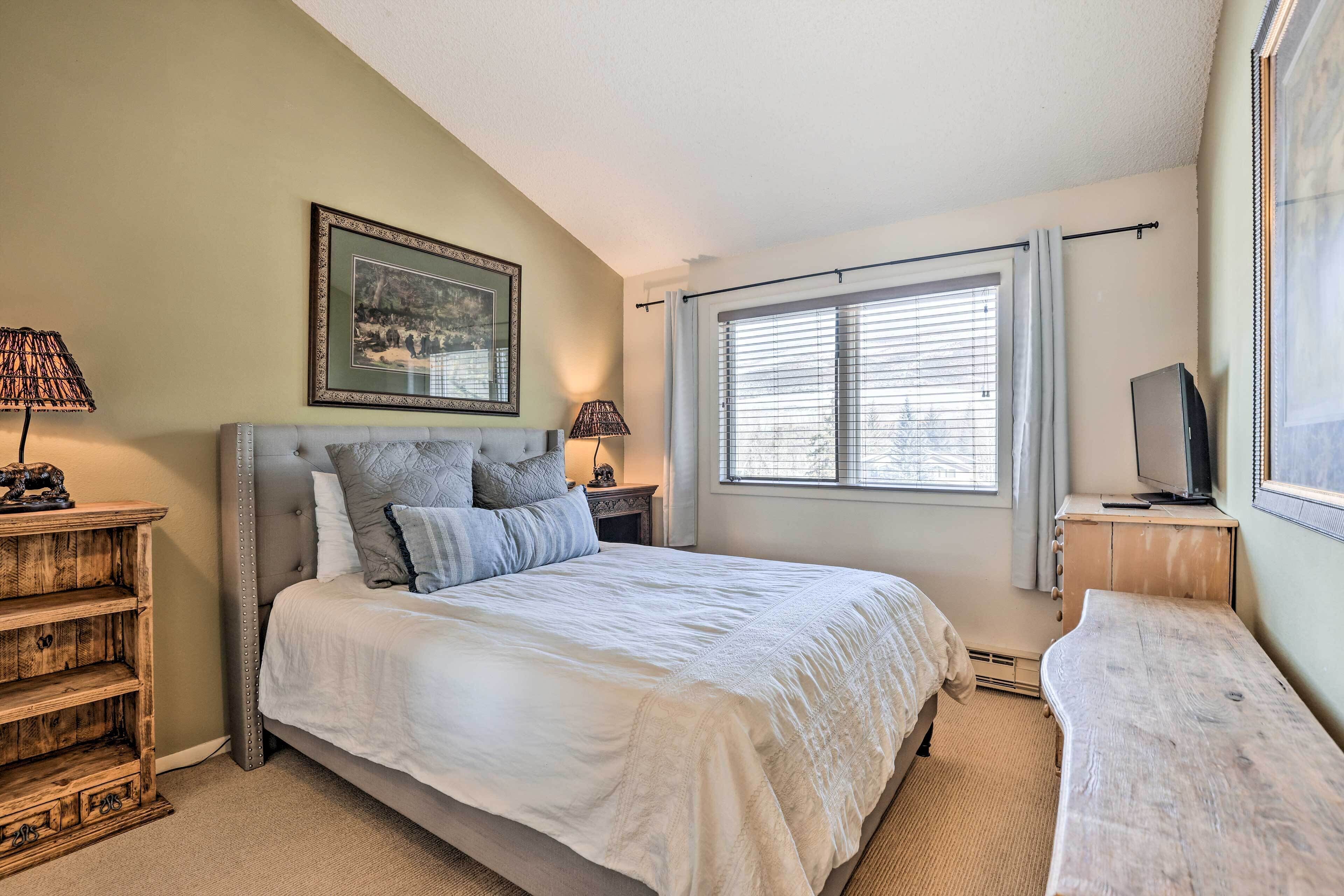 A bedroom with a bed, a nightstand, a dresser, a television, and a window with blinds.