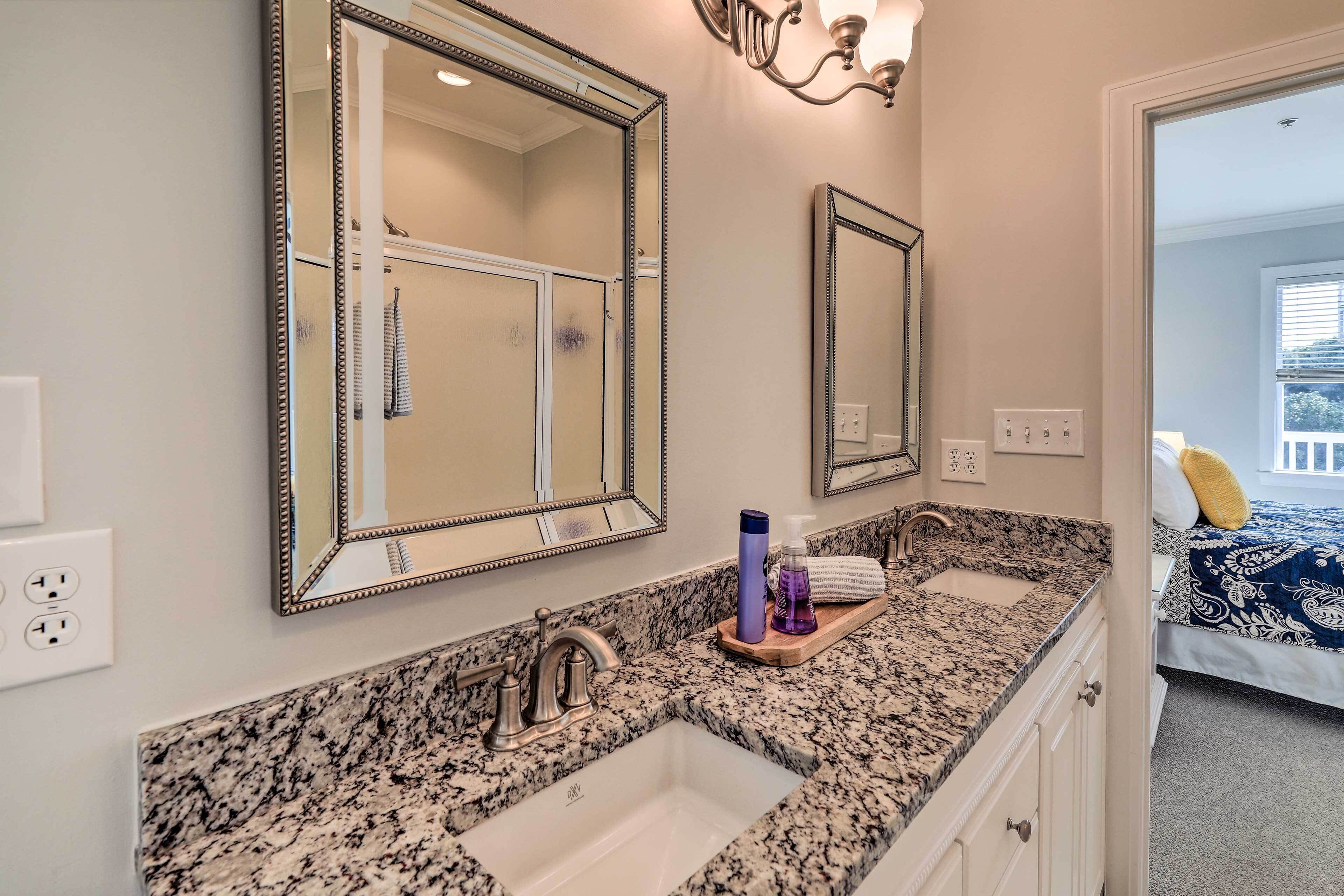A bathroom with a granite countertop, two sinks, and a large mirror. There is a towel rack and a bottle of hand soap on the countertop.