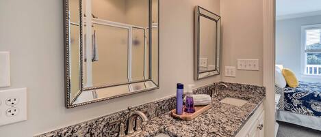 A bathroom with a granite countertop, two sinks, and a large mirror. There is a towel rack and a bottle of hand soap on the countertop.