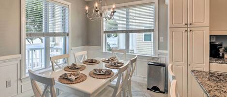A dining area with a white table set for six, surrounded by white chairs, a chandelier, and large windows with blinds.