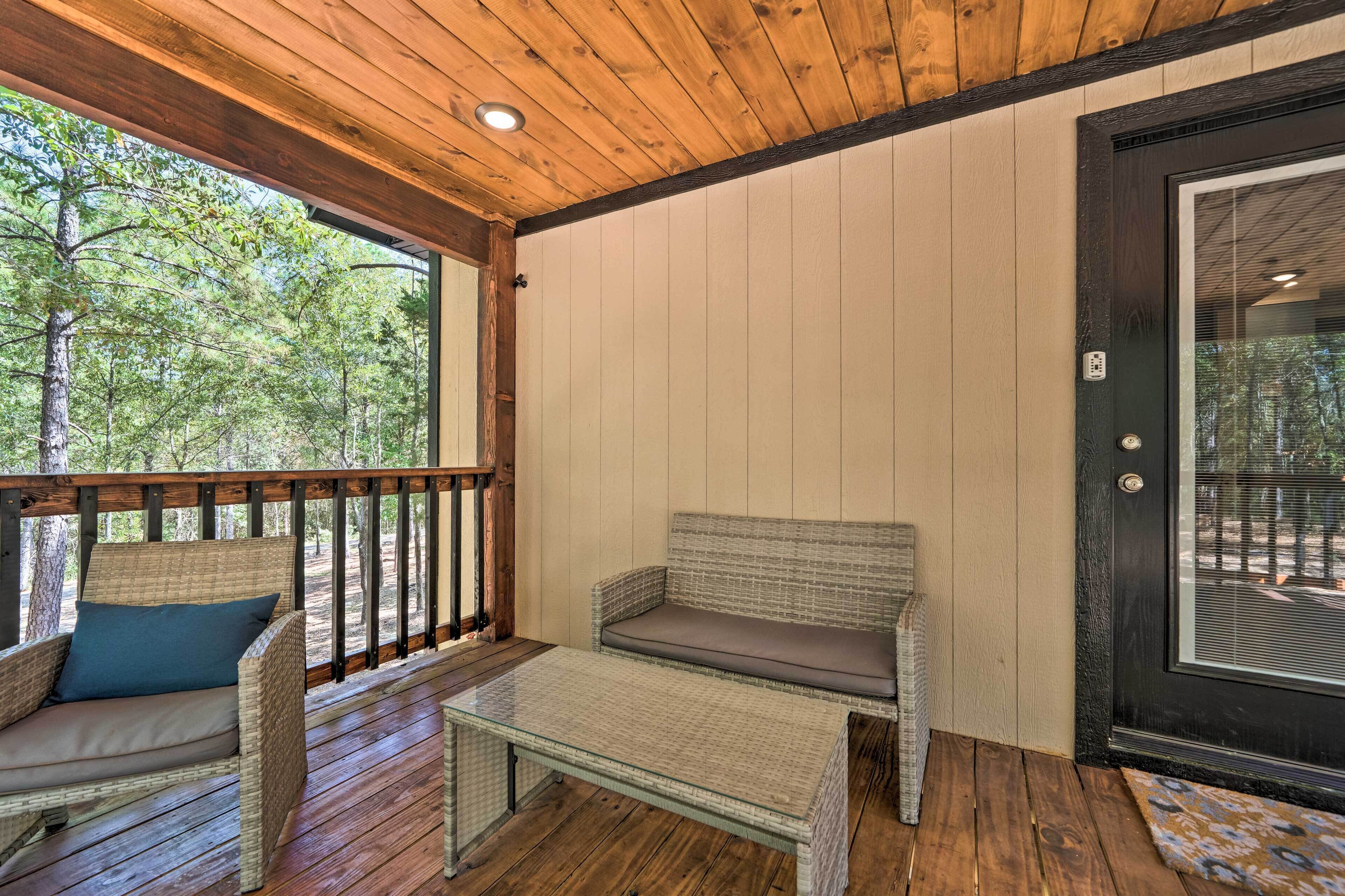 A covered porch with wicker furniture, a glass table, and a door leading to an outdoor area with trees.