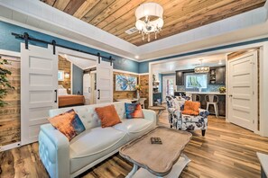 A spacious living room with a light blue sofa, a patterned armchair, and a wooden coffee table. A kitchen is visible in the background with a large window. - Modern Broken Bow Cabin, Near Lake + Wineries (Broken Bow)