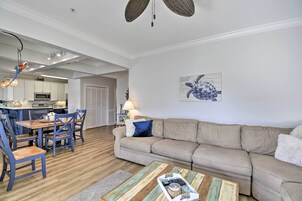 Una sala de estar moderna con un sofá color beige, una mesa de comedor de madera y sillas. Se ve una cocina con armarios blancos y un microondas al fondo.