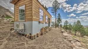 A wooden cabin with large windows, a small attached utility shed, and a dirt ground with scattered rocks.