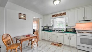 Une cuisine avec des meubles blancs, une table à manger avec des chaises, une cuisinière et une fenêtre avec des stores.