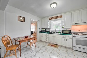 Une cuisine avec des meubles blancs, une table à manger avec des chaises, une cuisinière et une fenêtre avec des stores.