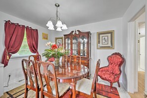 Une salle à manger avec une table en bois, six chaises, un fauteuil recouvert de tissu rouge, un lustre et un vase rempli de fleurs.
