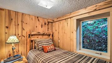 A wooden room with a bed, a window with a forest view, a lamp, and a small table.