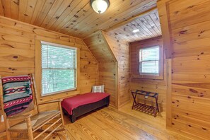 A cozy cabin room with wooden walls and floors, a bed with a red blanket, a rocking chair, and a small table. - Pet-friendly Cabin w/ Fireplace, Smoky Mtn Views (Bryson City)