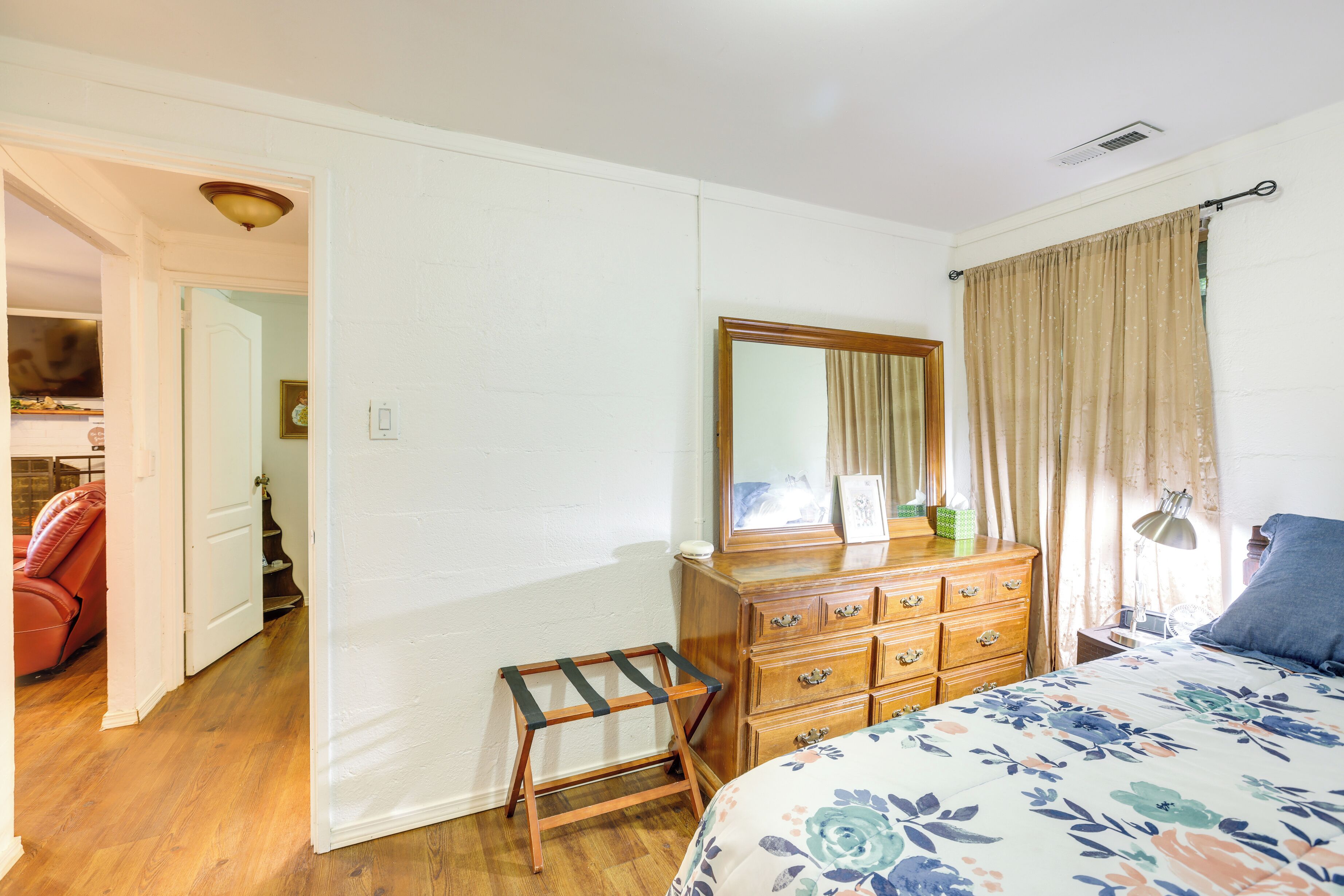 A bedroom with a wooden dresser, a bed with a floral bedspread, a mirror, and a window with curtains.