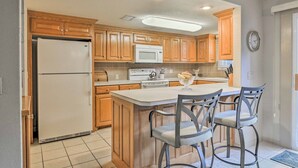 A kitchen with wooden cabinets, a white refrigerator, a microwave, and a stove. There are two bar stools at the island. - Lakefront Townhome w/ View in Hot Springs Village! (Hot Springs Village)