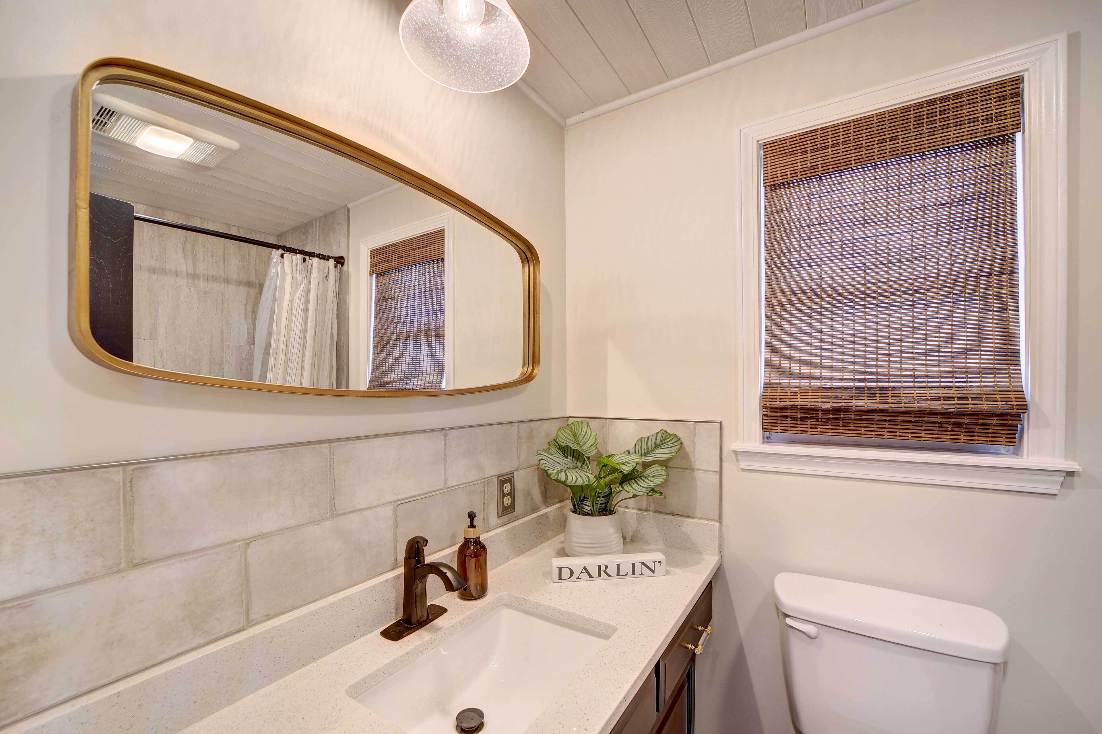 Une salle de bain avec un lavabo blanc, un miroir avec un cadre doré, des toilettes et une fenêtre avec des stores.