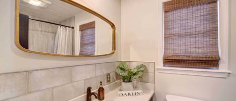 Une salle de bain avec un lavabo blanc, un miroir avec un cadre doré, des toilettes et une fenêtre avec des stores.