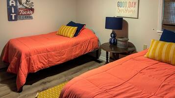 A room with two beds, a small round table with a lamp, a wall sign, and a window with blinds.
