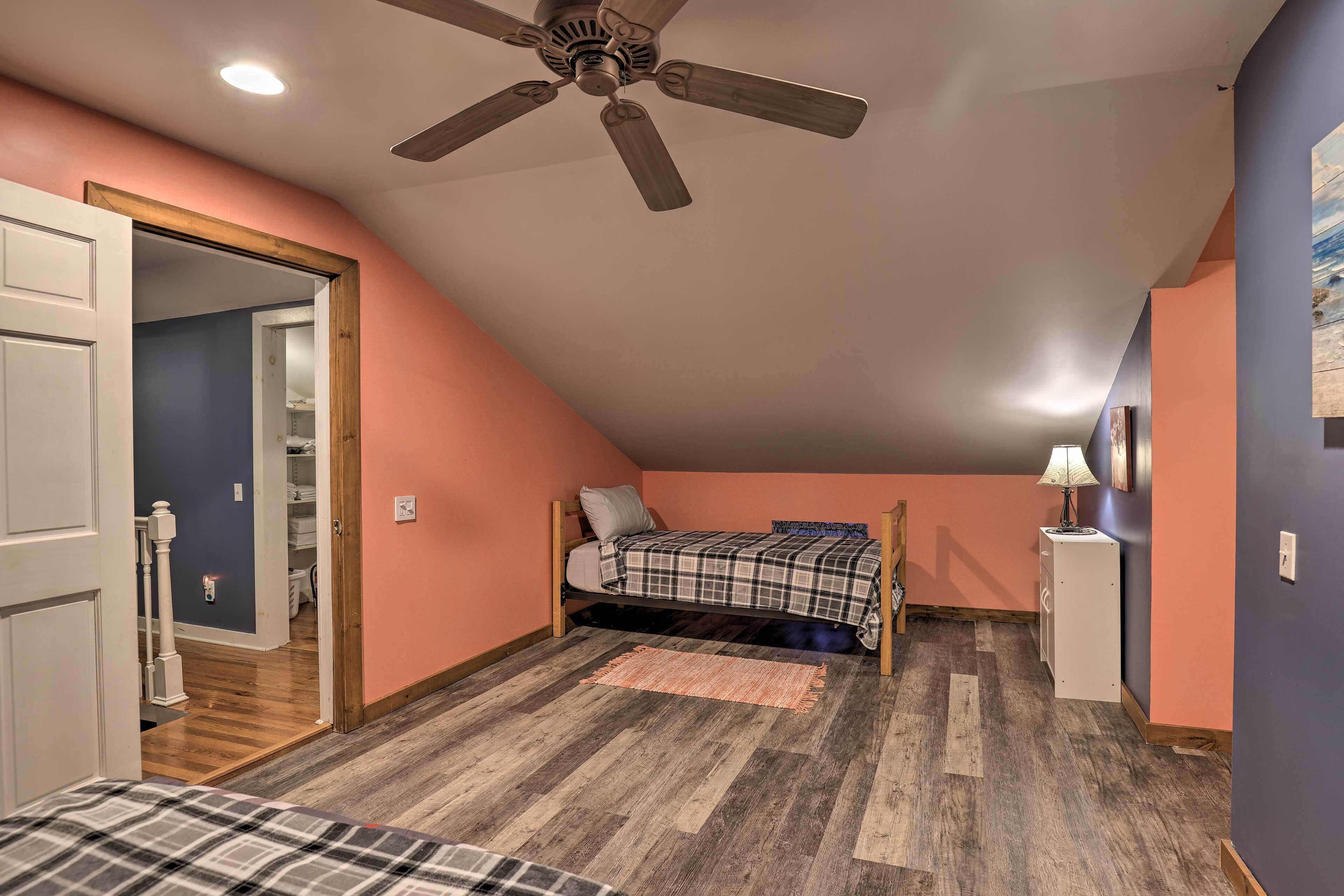 A bedroom with a sloped ceiling, a ceiling fan, a bed with a plaid bedspread, a bedside table with a lamp, and a wooden floor.