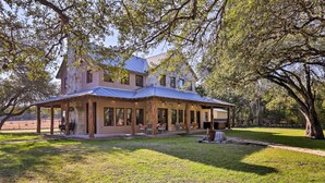 A large, two-story house with a covered patio area, surrounded by a well-maintained lawn and trees. - Riverfront Blanco Home w/ Shaded Porch + Hot Tub (Blanco)