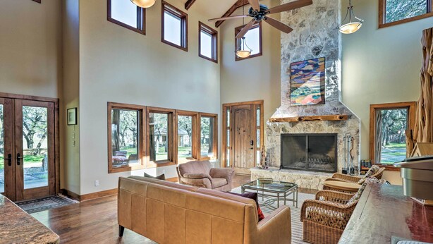 A spacious living room with a stone fireplace, large windows, and a high ceiling fan. - Riverfront Blanco Home w/ Shaded Porch + Hot Tub (Blanco)