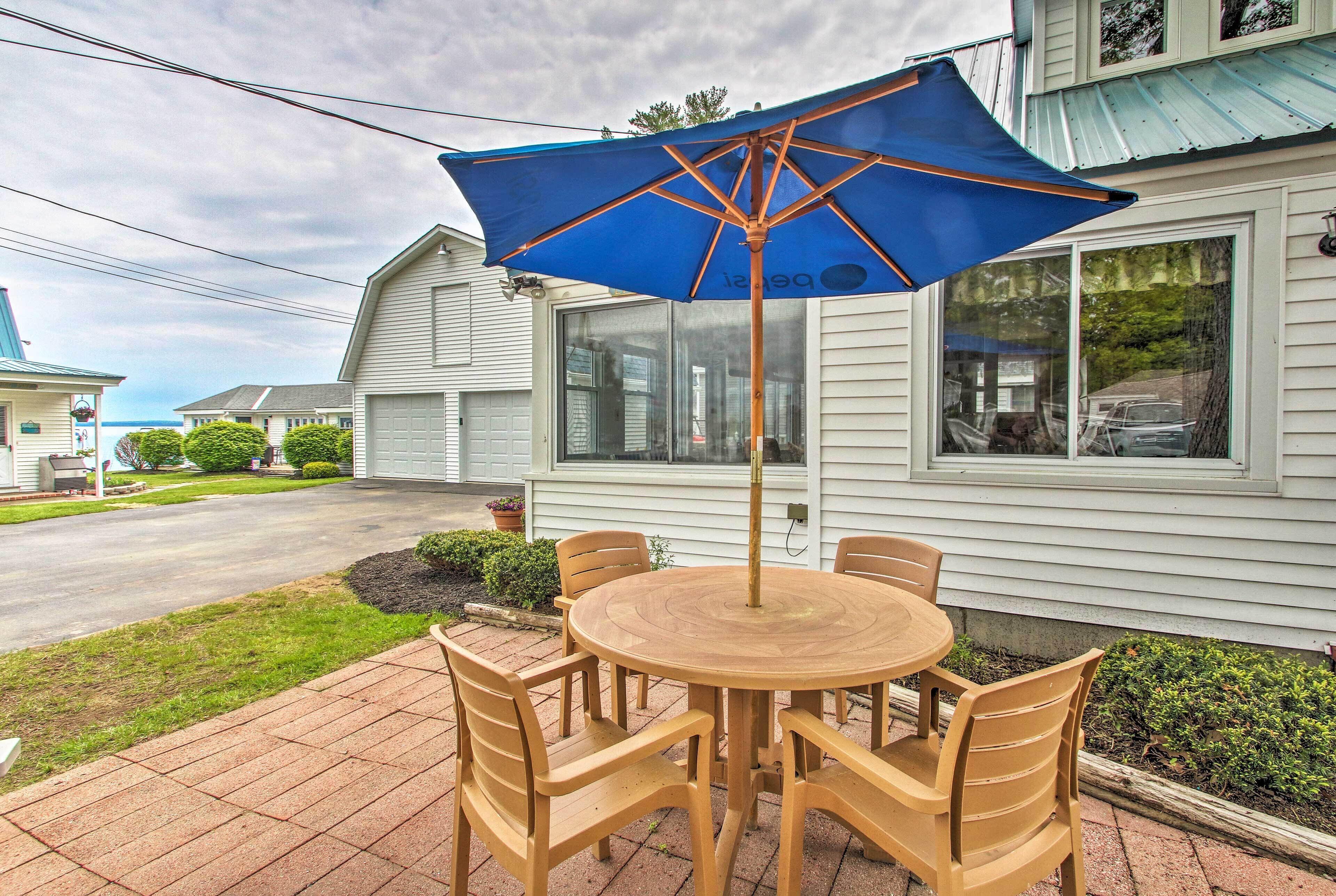 A patio with a round wooden table and chairs, a blue umbrella, and a house in the background.