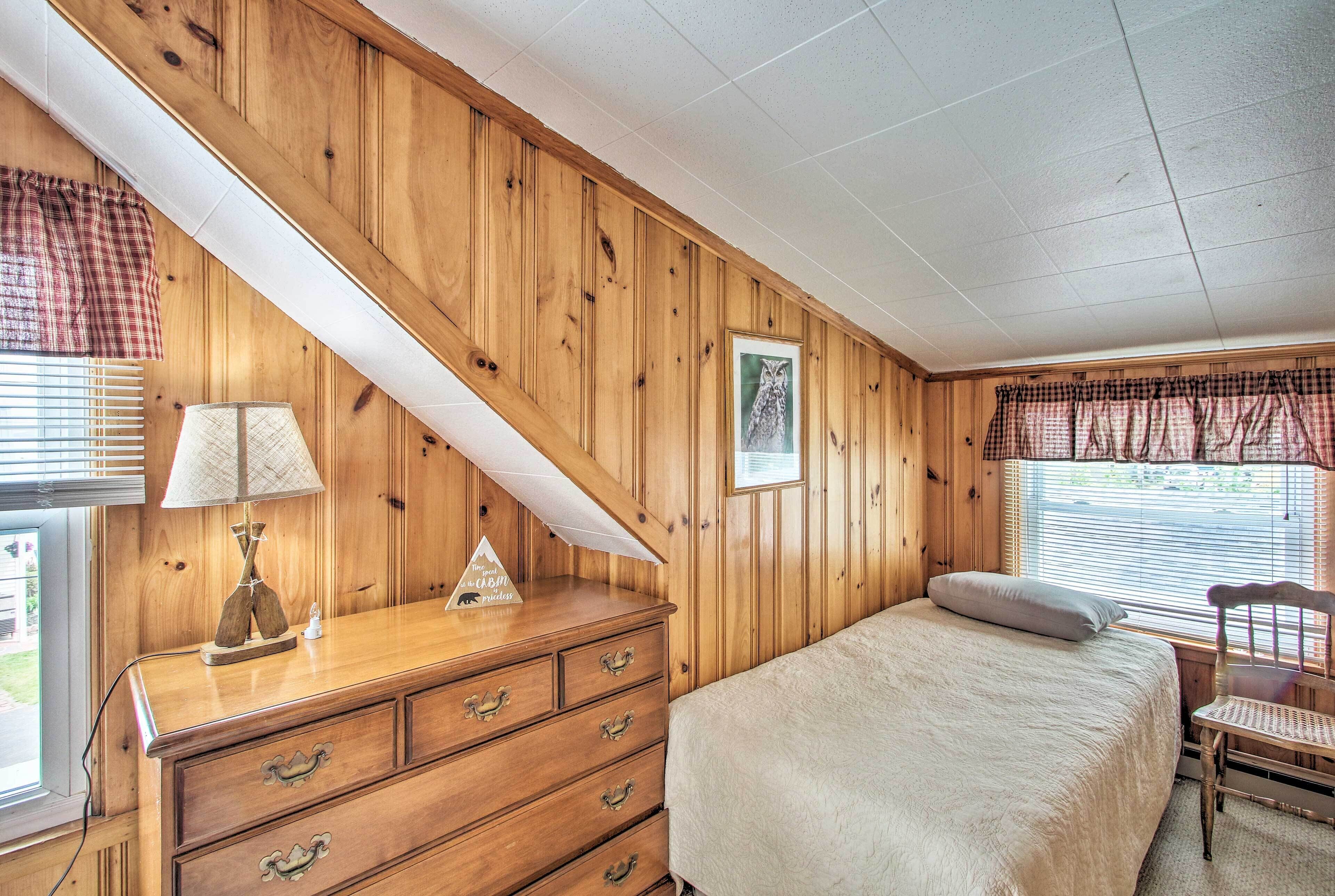 Une chambre avec un lambris en bois, un lit recouvert d’une couverture blanche, une commode à tiroirs, une lampe, un tableau encadré et une fenêtre avec des stores.