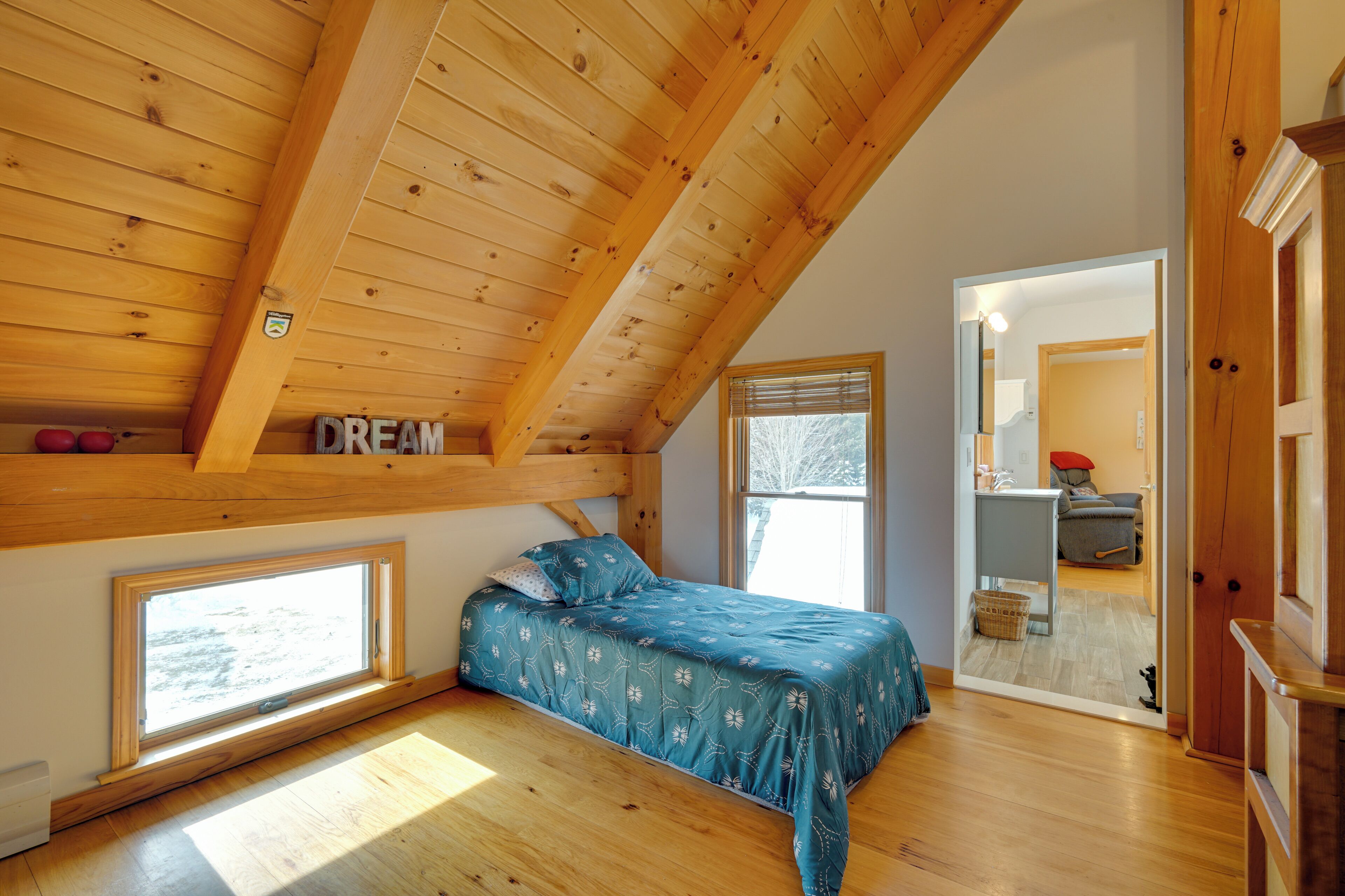 A bedroom with a sloped ceiling, wooden beams, a bed with a blue floral bedspread, and a window with a view of the outdoors.