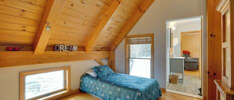 A bedroom with a sloped ceiling, wooden beams, a bed with a blue floral bedspread, and a window with a view of the outdoors.
