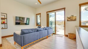 A modern living room with a blue sofa, wooden flooring, a ceiling fan, and large windows offering a view of mountains. - Modern Fraser Townhome w/ Private Hot Tub & Decks! (Fraser)