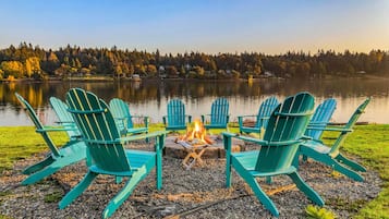 Eine Sitzgruppe am Seeufer mit vier grünen Adirondack-Stühlen, die um eine zentrale Feuerstelle angeordnet sind.