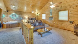 A cozy cabin interior with a wooden ceiling, a sofa, a coffee table, a fireplace, and a large window.