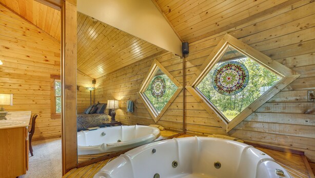 A wooden cabin with two hot tubs, a dining area, and a cozy seating area with a view through a stained glass window.