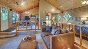 A spacious cabin with a high ceiling, wooden walls, a cozy living area with a sofa and coffee table, a dining area with a table and chairs, and a kitchen with a window.