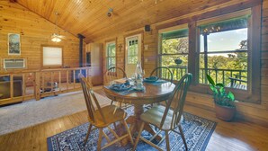 A wooden interior with a round dining table, chairs, a chandelier, and large windows with a view.