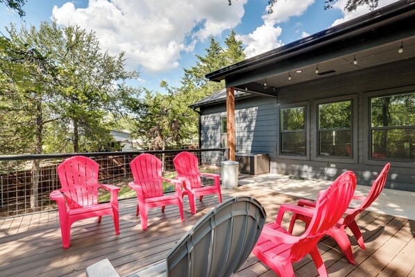 A deck with four red Adirondack chairs, a round table, and a large window overlooking a wooded area.