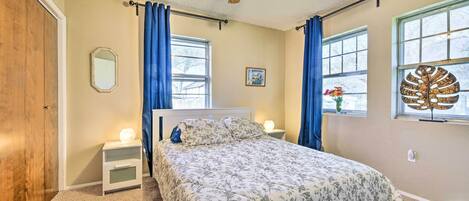 Un dormitorio con cama, ventilador de techo, dos ventanas con cortinas, una mesita de noche y un espejo montado en la pared.