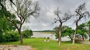 A grassy area with a stone wall, trees, and a view of a lake. - Sunny Lakefront Family Paradise w/ Deck & 2 Kayaks (Granbury)