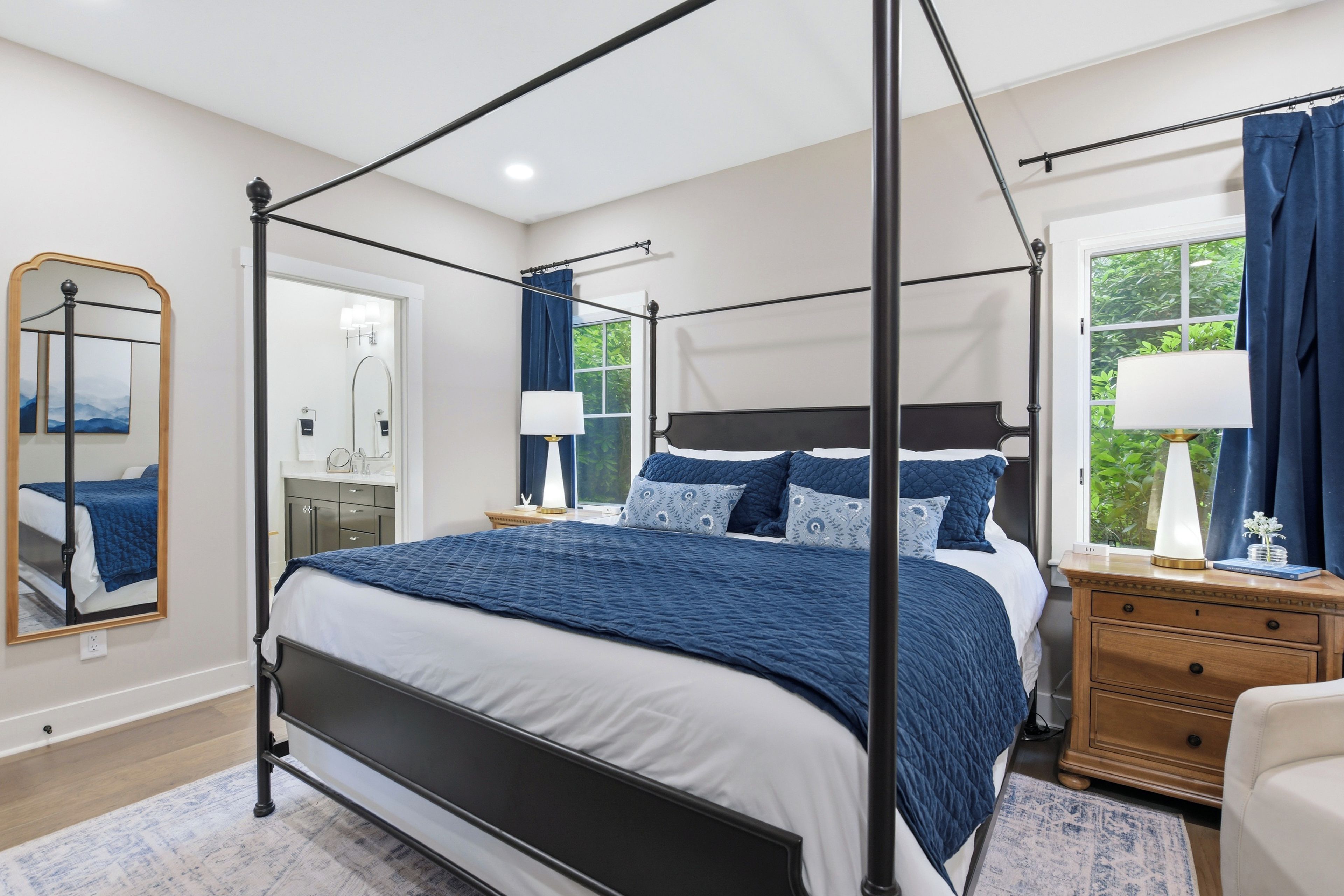 A four-poster bed with a white and blue bedspread, a wooden nightstand, a mirror, and a window with curtains.