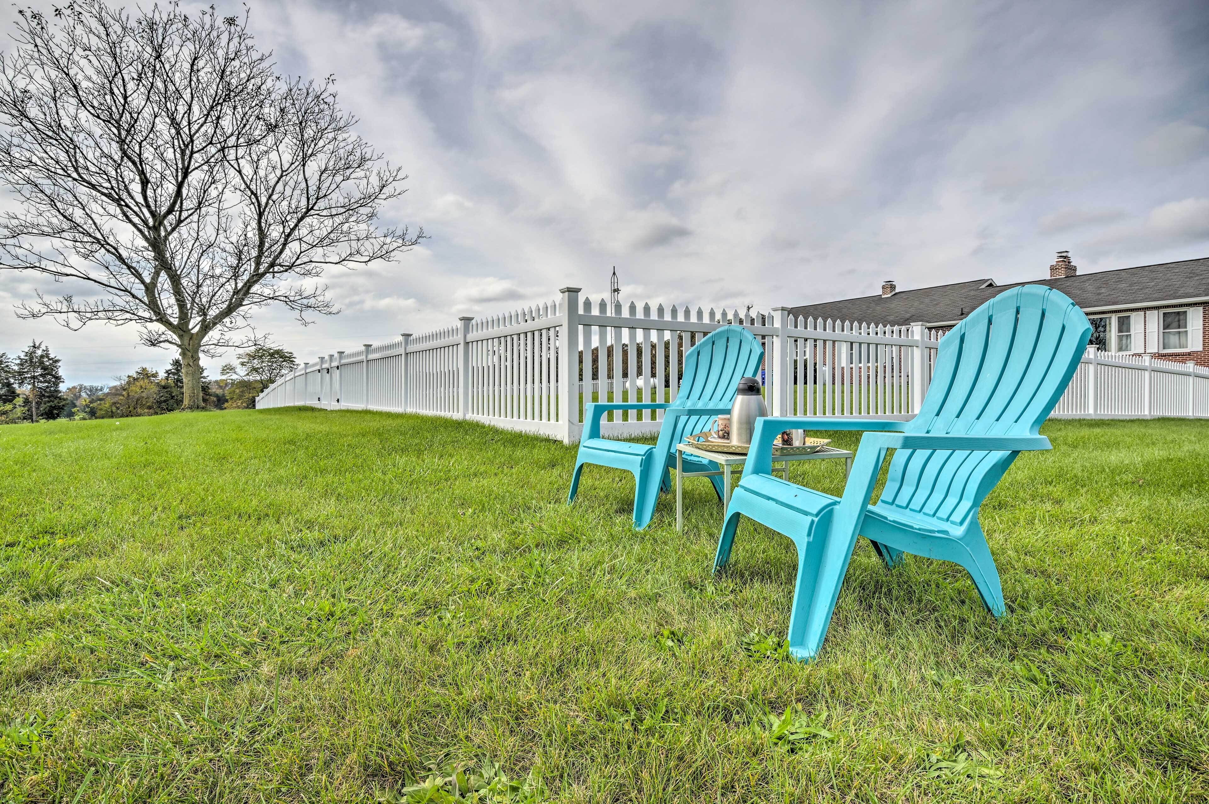 A grassy lawn with two blue Adirondack chairs and a white picket fence.