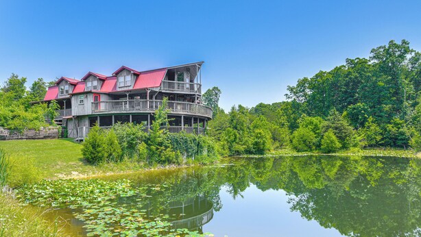 A large, multi-story wooden house with a red roof, surrounded by lush greenery and a calm lake. - Strawberry Plains Cabin: Private Pond & Game Room! (Kodak)