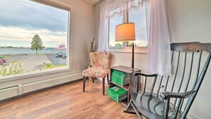 A room with a large window overlooking a body of water, a chair, a lamp, and a small table with green boxes.