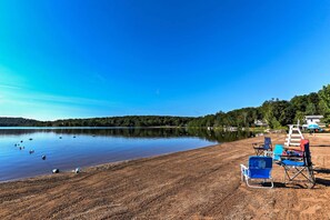 A beach with a clear sky, a calm lake, and several beach chairs and umbrellas. - Arrowhead Lake Amenities: Quaint Home w/ Fireplace (Pocono Lake)