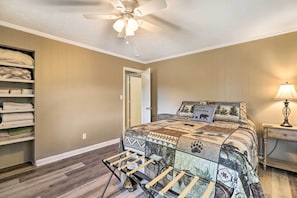 Ein Schlafzimmer mit einem Bett, einem Deckenventilator, einem Nachttisch, einer Lampe, einem Regal mit zusammengefalteten Decken und einer Tür zu einem anderen Raum.